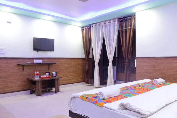 Habitación de hotel con cama, televisor montado en la pared, una mesita con hervidor y bandeja, y cortinas.