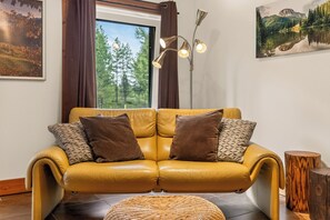A yellow leather sofa with two brown pillows, a woven ottoman, a tall floor lamp, and a large window with curtains.