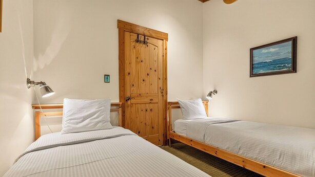 Quarto com duas camas de solteiro, uma porta de madeira, um quadro na parede e uma luminária pequena.