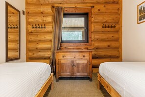 Quarto com duas camas, paredes de madeira, uma janela com cortinas e um armário pequeno.