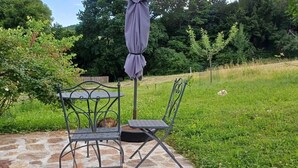 Een terras buiten met een metalen tafel en stoelen, een stenen vloer en een gazon met bomen op de achtergrond.