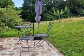 Eine Terrasse im Freien mit einem Metalltisch und -stühlen, einem Steinfußboden und einem Rasenbereich mit Bäumen im Hintergrund.
