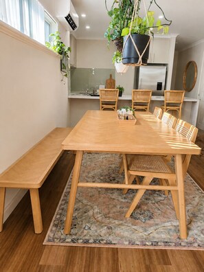 A dining area with a wooden table and benches, a hanging plant, and a kitchen in the background. - Yarri House- walk to shops, estuary and beach! (Falcon)