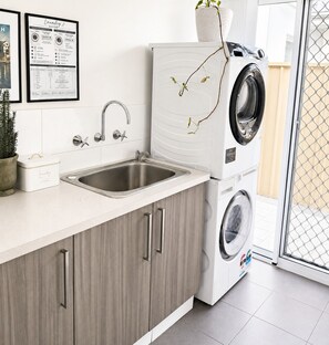 A modern laundry room with a sink, a washing machine, and a dryer stacked on top of each other. - Yarri House- walk to shops, estuary and beach! (Falcon)