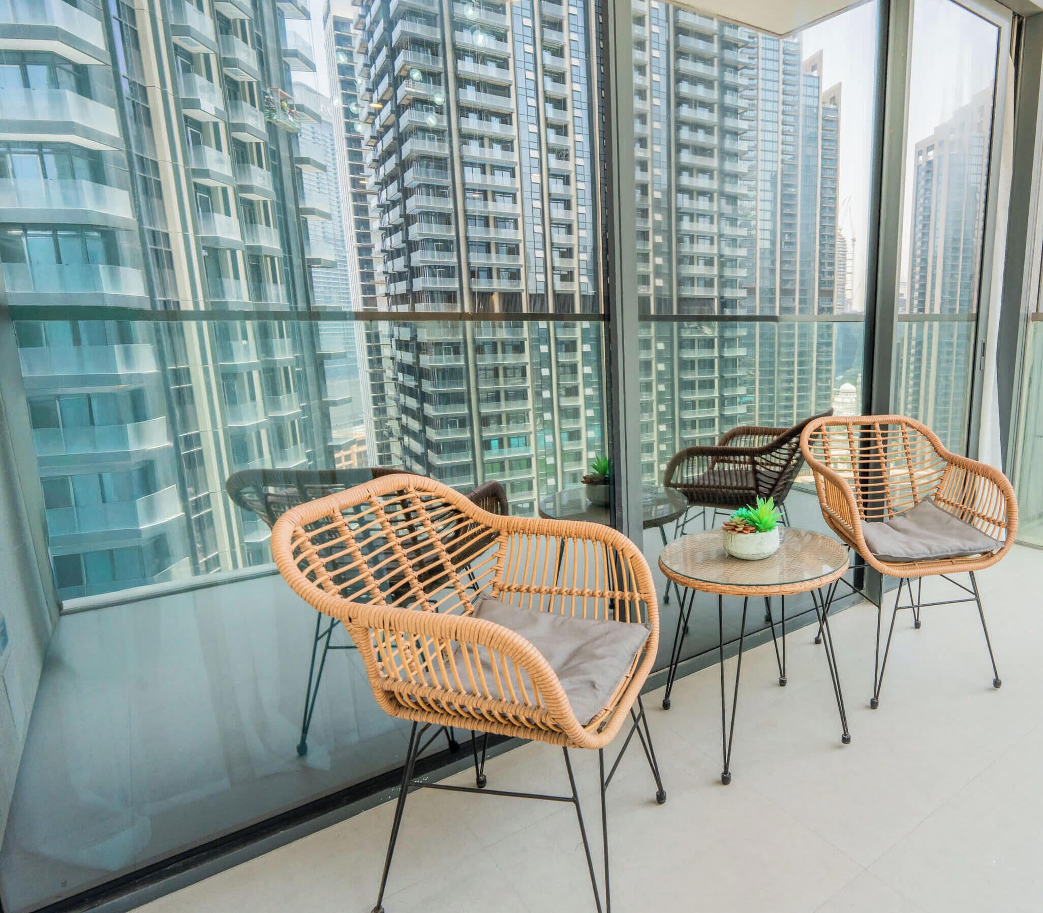 A balcony with wicker chairs and a small table, overlooking a cityscape with high-rise buildings.