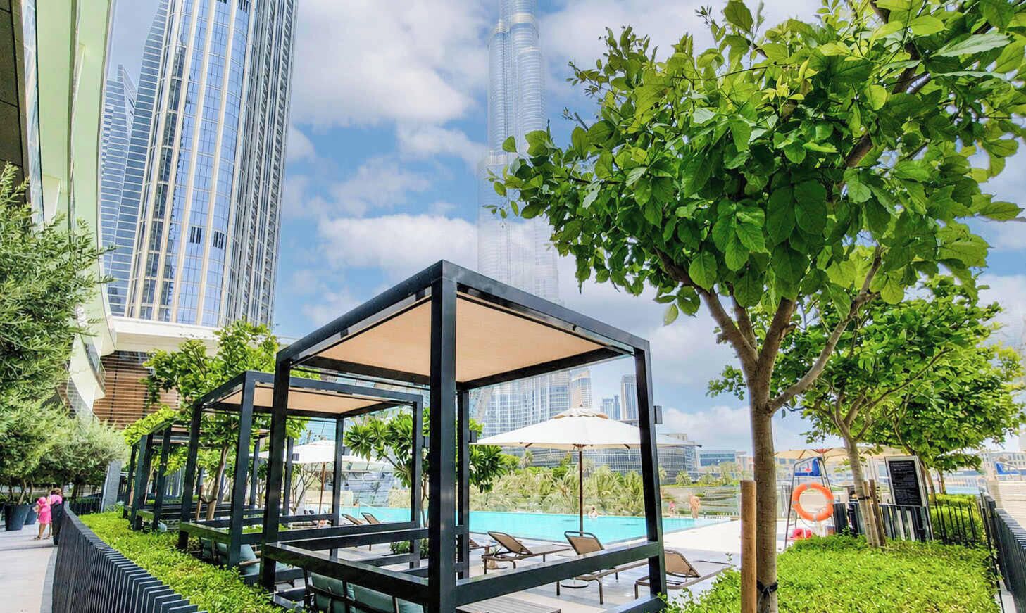 A rooftop pool area with lounge chairs and a glass-enclosed seating area, surrounded by greenery and modern buildings in the background.