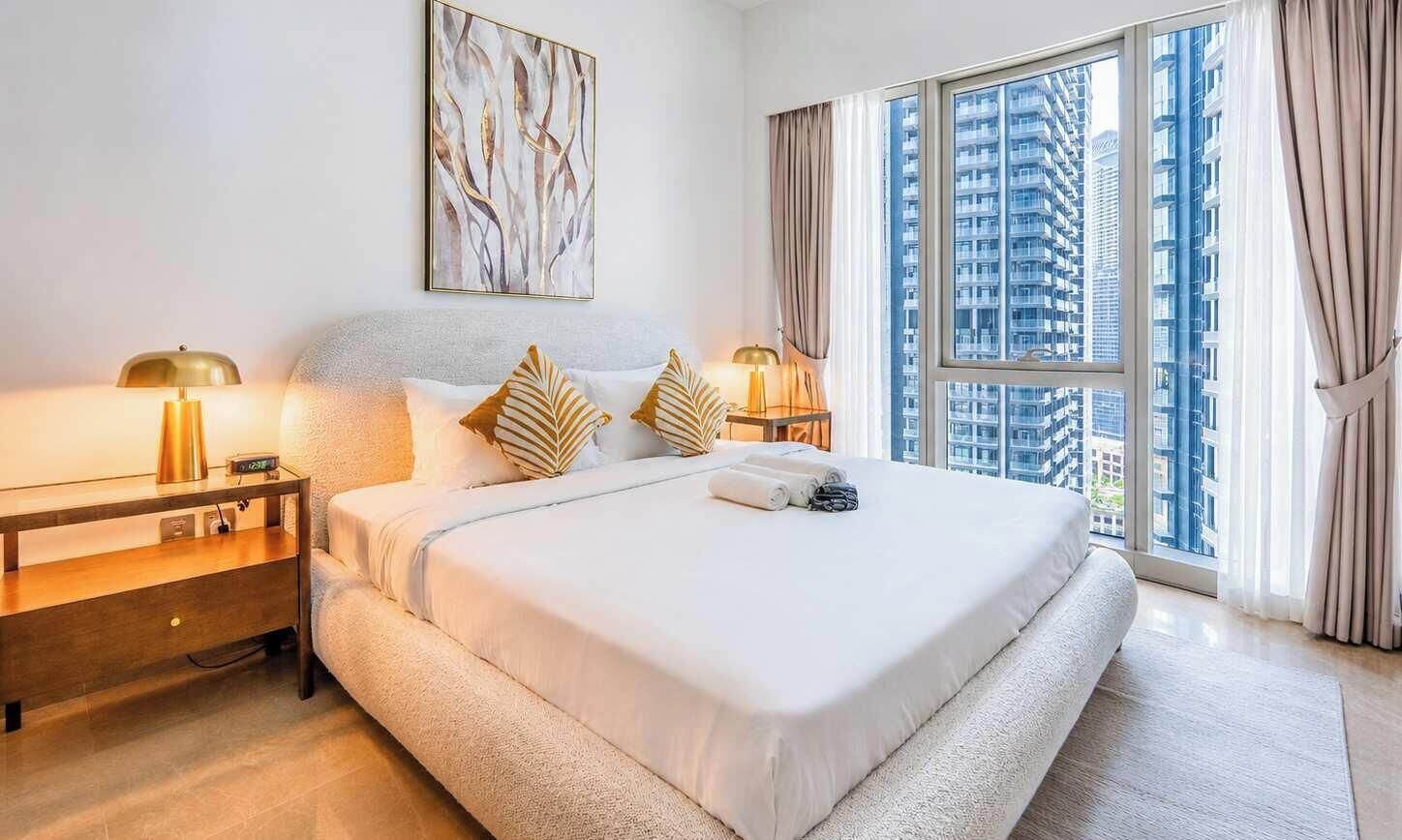A hotel room with a large bed, two bedside tables with lamps, a view of high-rise buildings, and a large window with curtains.