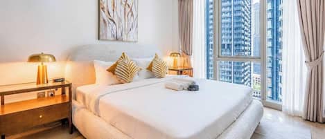 Habitación de hotel con una cama grande, dos mesitas de noche con lámparas, vistas a edificios altos y un ventanal con cortinas.