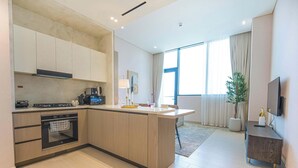 A modern kitchen with a built-in oven and a coffee machine, a dining area with a table and chairs, a large window with curtains, and a flat-screen TV on a stand. - Modern 1BR, Binghatti Royale, JVC (Dubai)