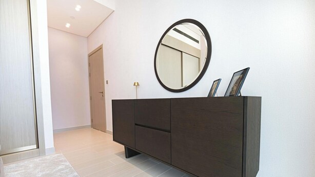 A hallway with a dark wooden cabinet, a round mirror, and a door. - Modern 1BR, Binghatti Royale, JVC (Dubai)