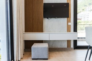 A modern living room with a flat-screen TV mounted on a wall, a built-in bench, and a view of a building through a large window. - Bella Vista Balcony | 1BR Apartment by PikHost (Vlorë)
