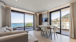 A modern living room with a sofa, dining table, and chairs, large windows with a view of the sea, and a flat-screen TV mounted on the wall.