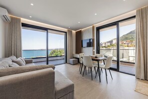 A modern living room with a sofa, dining table, and chairs, large windows with a view of the sea, and a flat-screen TV mounted on the wall. - Bella Vista Balcony | 1BR Apartment by PikHost (Vlorë)