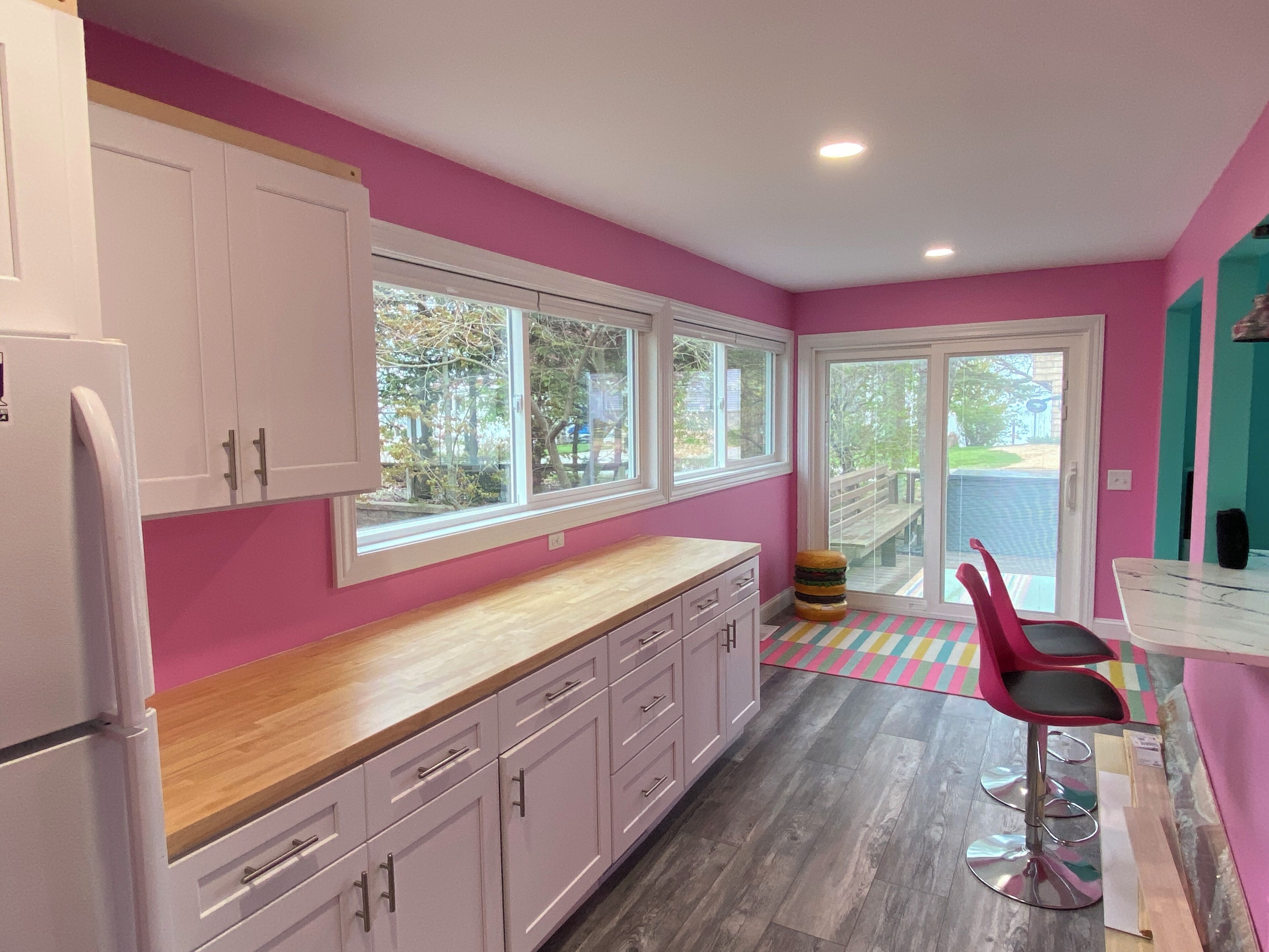 A kitchen with pink walls, white cabinets, and a wooden countertop. There is a window with a view of trees and a door leading outside.
