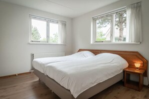 Een slaapkamer met een bed, twee ramen, een nachtkastje met een lamp en een radiator.