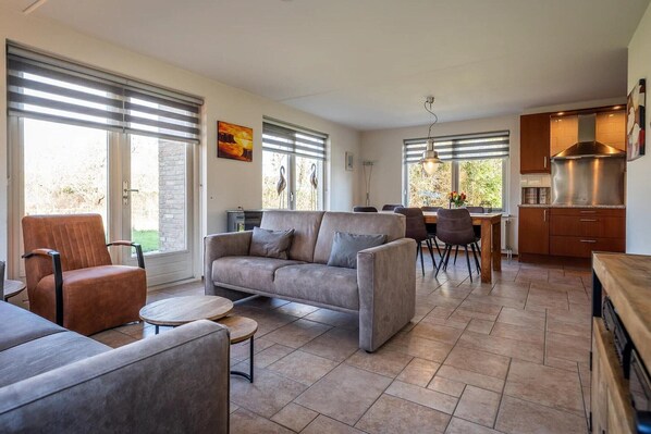 A modern living room with a sofa, armchairs, and a dining area with a table and chairs. There is a kitchen with a stove and cabinets.