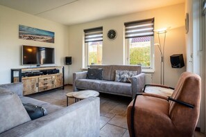 A modern living room with a grey sofa, a brown armchair, a flat-screen TV, a wooden cabinet, and a large window with blinds.