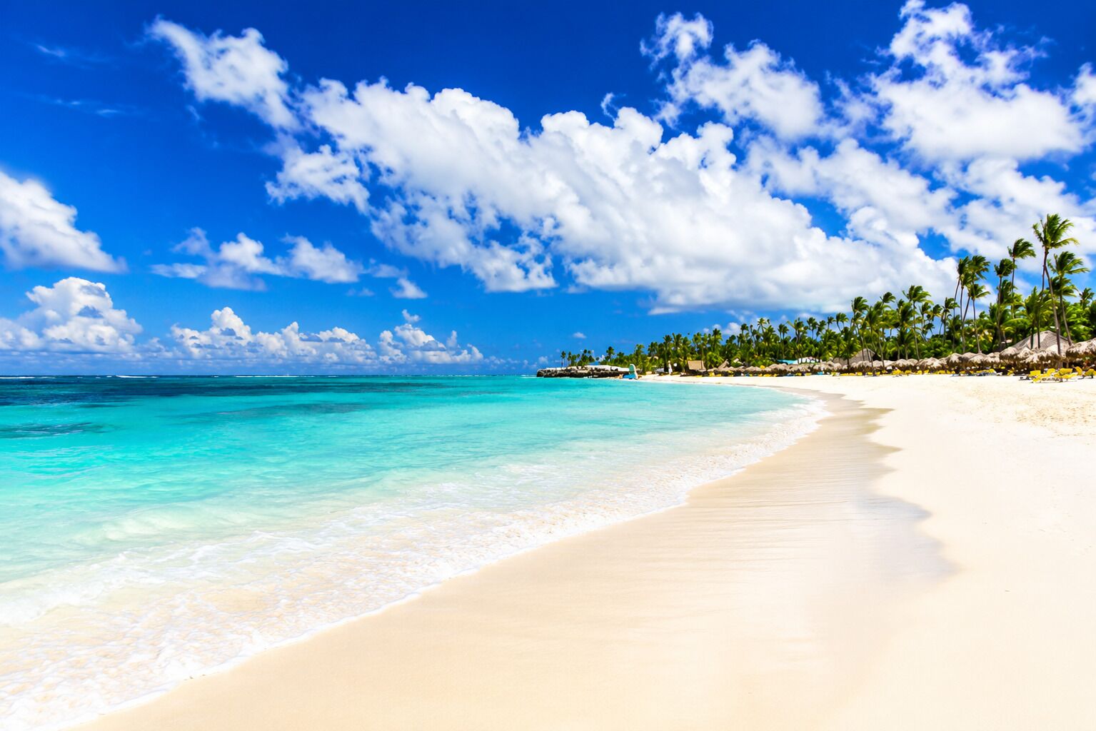 Une plage à l’eau turquoise et limpide, au sable blanc, bordée d’une rangée de palmiers sous un ciel bleu parsemé de nuages.