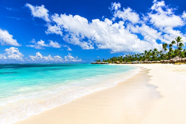 Une plage à l’eau turquoise et limpide, au sable blanc, bordée d’une rangée de palmiers sous un ciel bleu parsemé de nuages.