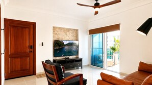 Un soggiorno con televisione, ventilatore a soffitto, una porta in legno e vista su una zona esterna attraverso una porta in vetro.