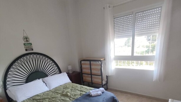 Quarto com cama, mesa de cabeceira, prateleira na parede, janela com persianas e ventoinha no teto.