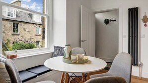 A cozy room with a round table, chairs, a window with a view of a stone building, and a radiator on the wall. - Stylish 2-Bed Villa · Central Edinburgh (Edinburgh)
