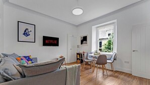 A modern living room with a grey sofa, a dining area with a table and chairs, a bookshelf, and a window with a view of greenery. - Stylish 2-Bed Villa · Central Edinburgh (Edinburgh)