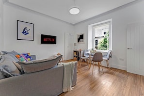 Ein modernes Wohnzimmer mit einem grauen Sofa, einem Essbereich mit Tisch und Stühlen, einem Bücherregal und einem Fenster mit Blick auf Grünflächen.