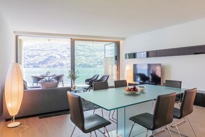 Ein moderner Essbereich mit einem Glastisch, schwarzen Stühlen, einem Fernseher und Blick auf eine Berglandschaft.