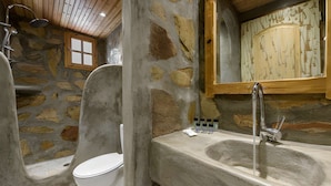 A stone-walled bathroom with a white toilet, a bathtub, and a shower. - Kyanee (Adamantas)