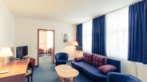 Habitación de hotel con sofá, sillas, escritorio con televisor, una mesita y ventana con cortinas.