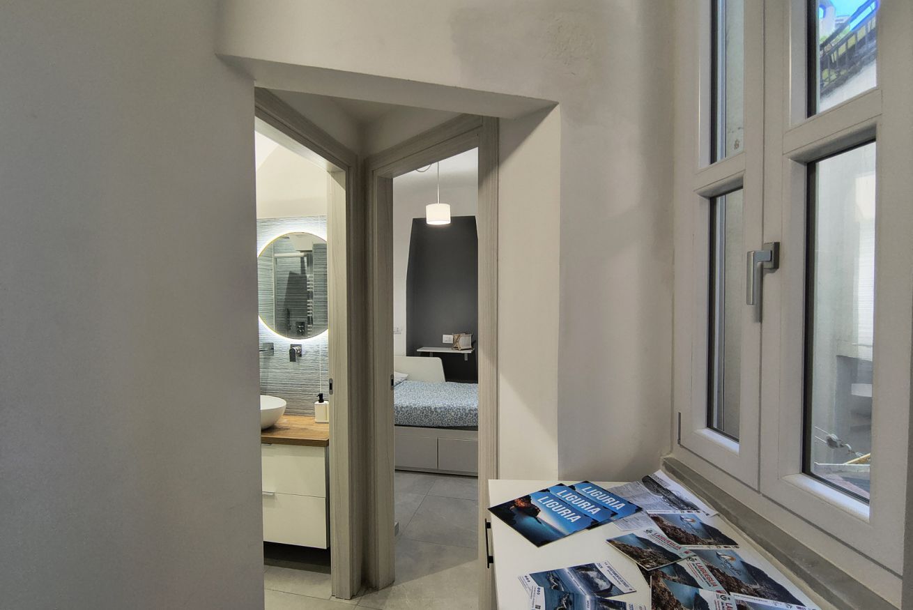 A narrow hallway leading to a bedroom with a bed, a bedside table with magazines, and a window with a view of the outside.