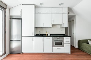 A modern kitchen with white cabinets, a stainless steel refrigerator, a built-in oven, and a green sofa in the adjacent living area. - Charming penthouse in a prime location (Madrid)