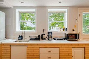 A modern kitchen with a sink, coffee maker, and microwave. There are two windows with a view of greenery outside. - Modern Studio Suite Near DC with Full Kitchen (Arlington)