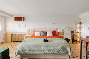 Quarto de hotel com cama, radiador, cadeira, mesa e prateleira com objetos decorativos.