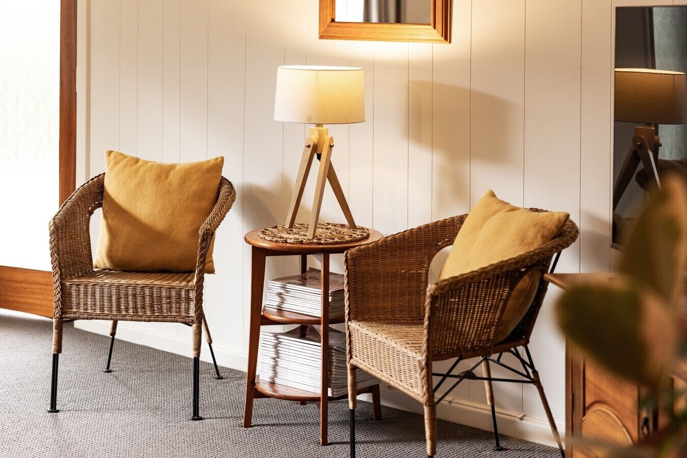 A room with two wicker chairs, a small round table with a lamp, and a mirror on the wall.