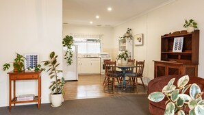 A living room with a dining area, a kitchen in the background, and a bookshelf with plants.