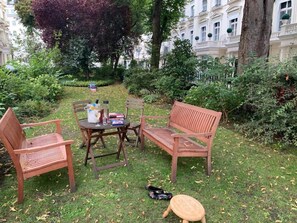 Ein Sitzbereich im Freien mit zwei Holzstühlen und einer Bank, einem runden Tisch mit einem Getränk und einem Buch sowie einem Paar Schuhe am Boden.