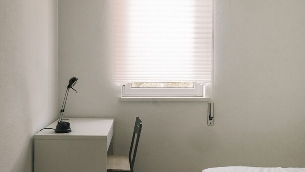 Ein Zimmer mit Schreibtisch, Stuhl, einem Fenster mit Jalousien und einem an der Wand befestigten Lichtschalter.
