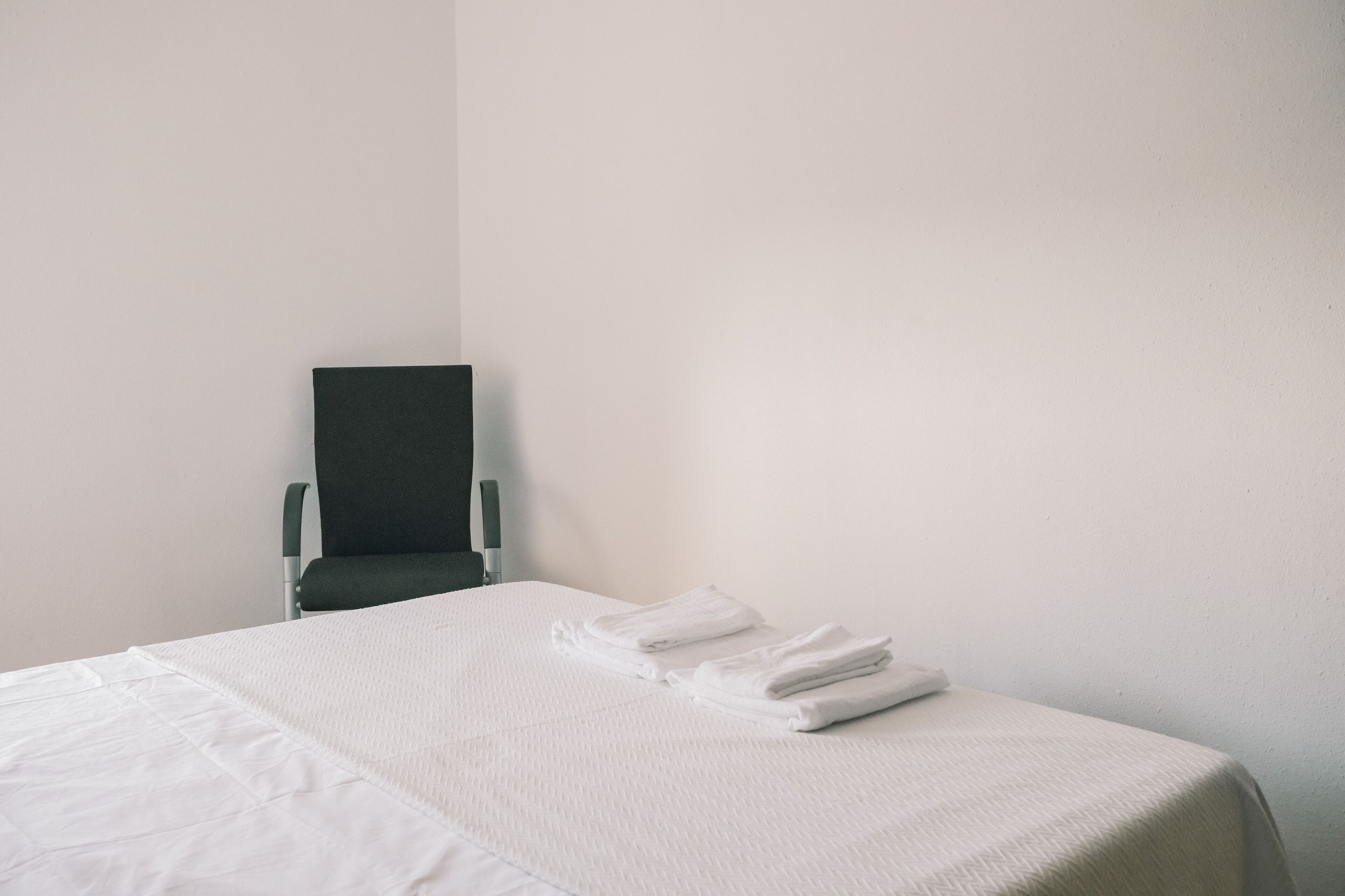Ein ordentlich gemachtes Bett mit weißen Leinen und gefalteten Handtüchern, ein Stuhl und eine schlichte Wand.