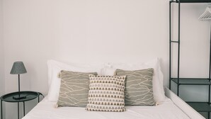 Ein Bett mit weißer Bettwäsche, zwei gemusterte Kissen, ein kleiner runder Tisch mit Lampe und ein Metallregal.