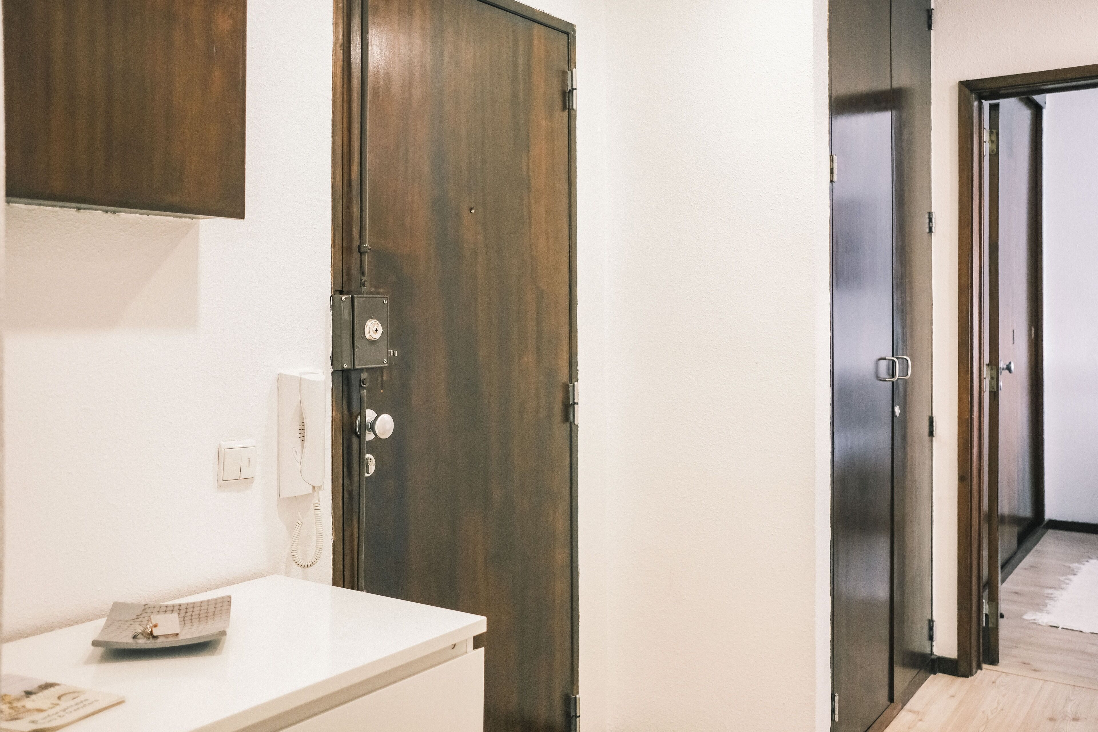 Un couloir avec une porte en bois, un mur blanc et un comptoir blanc sur lequel se trouvent un téléphone et un petit objet.