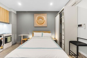 Ett modernt hotellrum med en säng, sängbord, ett konstverk på väggen, en garderob och ett litet kök med tvättmaskin.