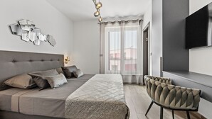 Ein modernes Schlafzimmer mit einem großen Bett, einer Kopfteil, einem Nachttisch, einem Sessel, einem Fernseher und einem Fenster mit Vorhängen.