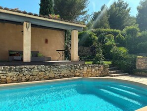 A swimming pool with a stone wall and a covered seating area with cushions. - Charmante Maison Provence 3Chambres piscine extérieure LANCON PROVENCE (Lançon-Provence)