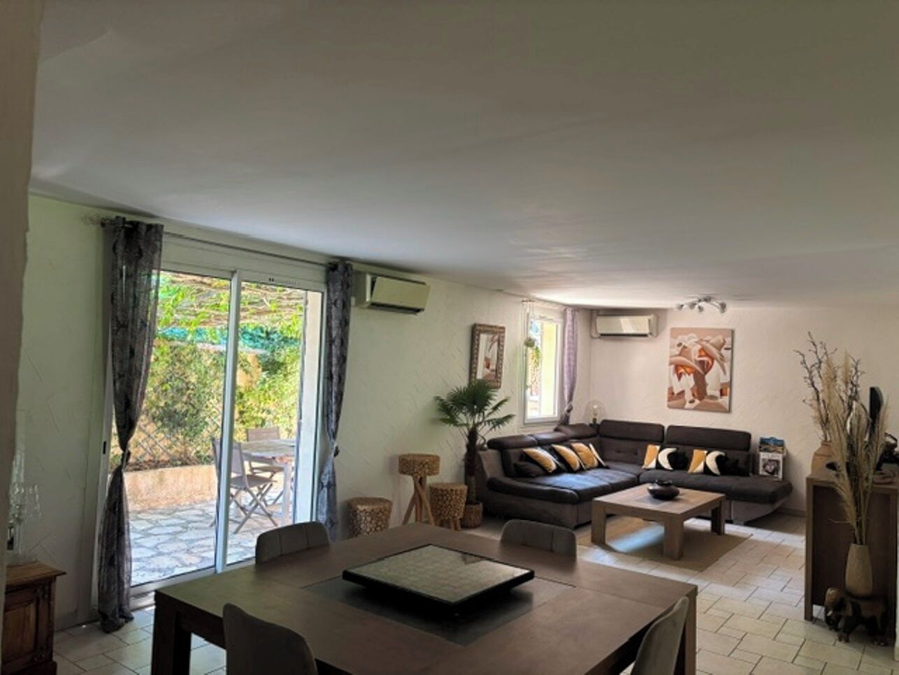 Ein Wohnzimmer mit einer Couch, einem Couchtisch und einem Esstisch. Ein großes Schiebeglastür führt zu einem Außenbereich.
