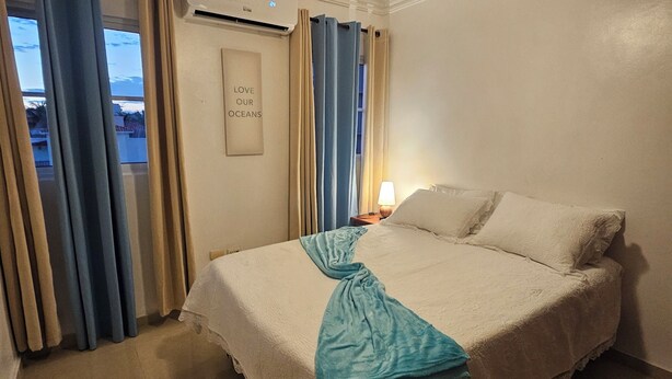 Quarto de hotel com cama, cortinas, ventoinha no teto, mesa de cabeceira e parede com mensagem emoldurada.