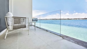 Un balcón con una silla blanca, una mesa pequeña y una barandilla de vidrio con vista al mar azul y despejado.