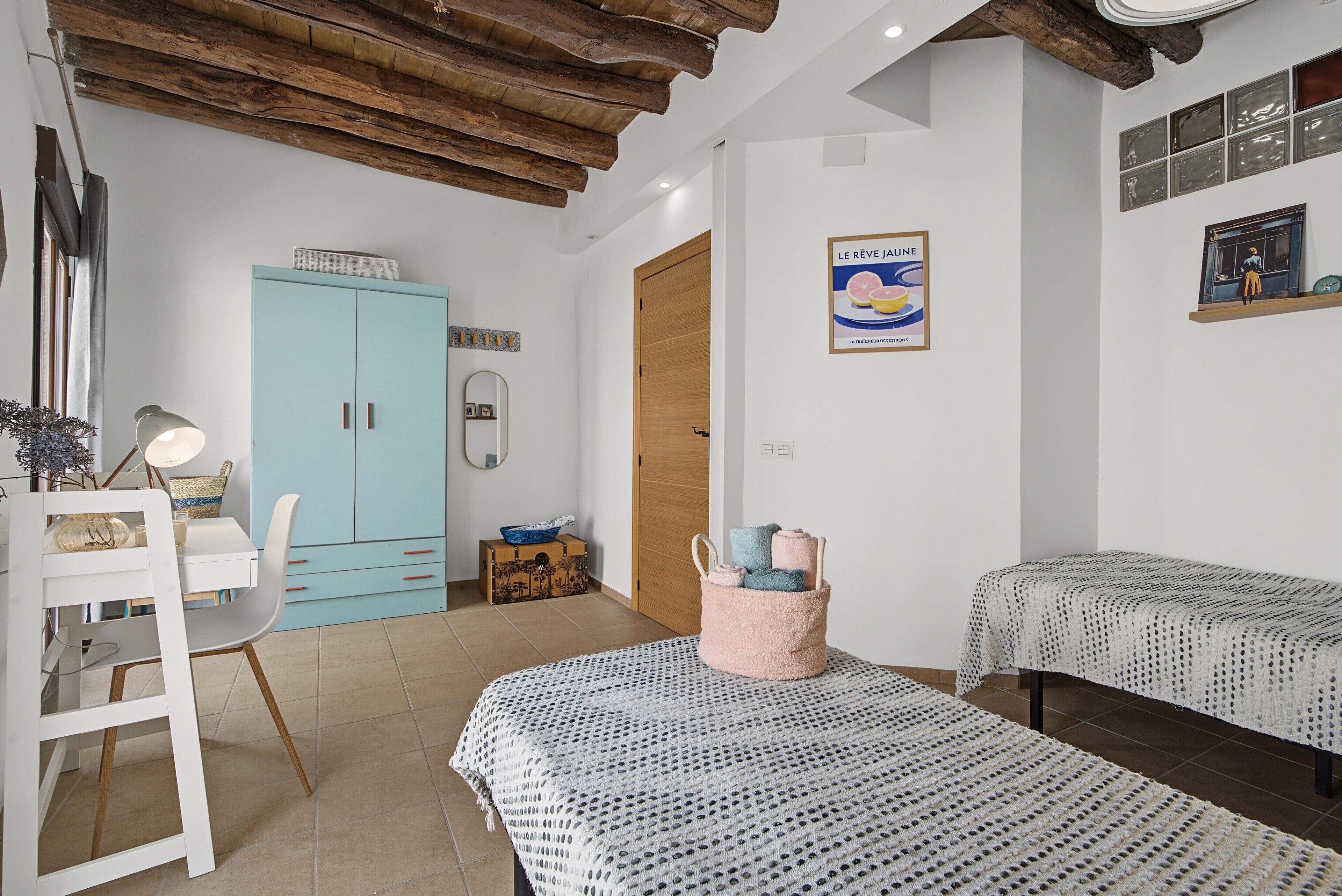 Ein Zimmer mit Holzboden, einem hellblauen Schrank, einem weißen Schreibtisch mit Lampe, zwei Betten mit gemusterten Betttextilien und einem Poster an der Wand.
