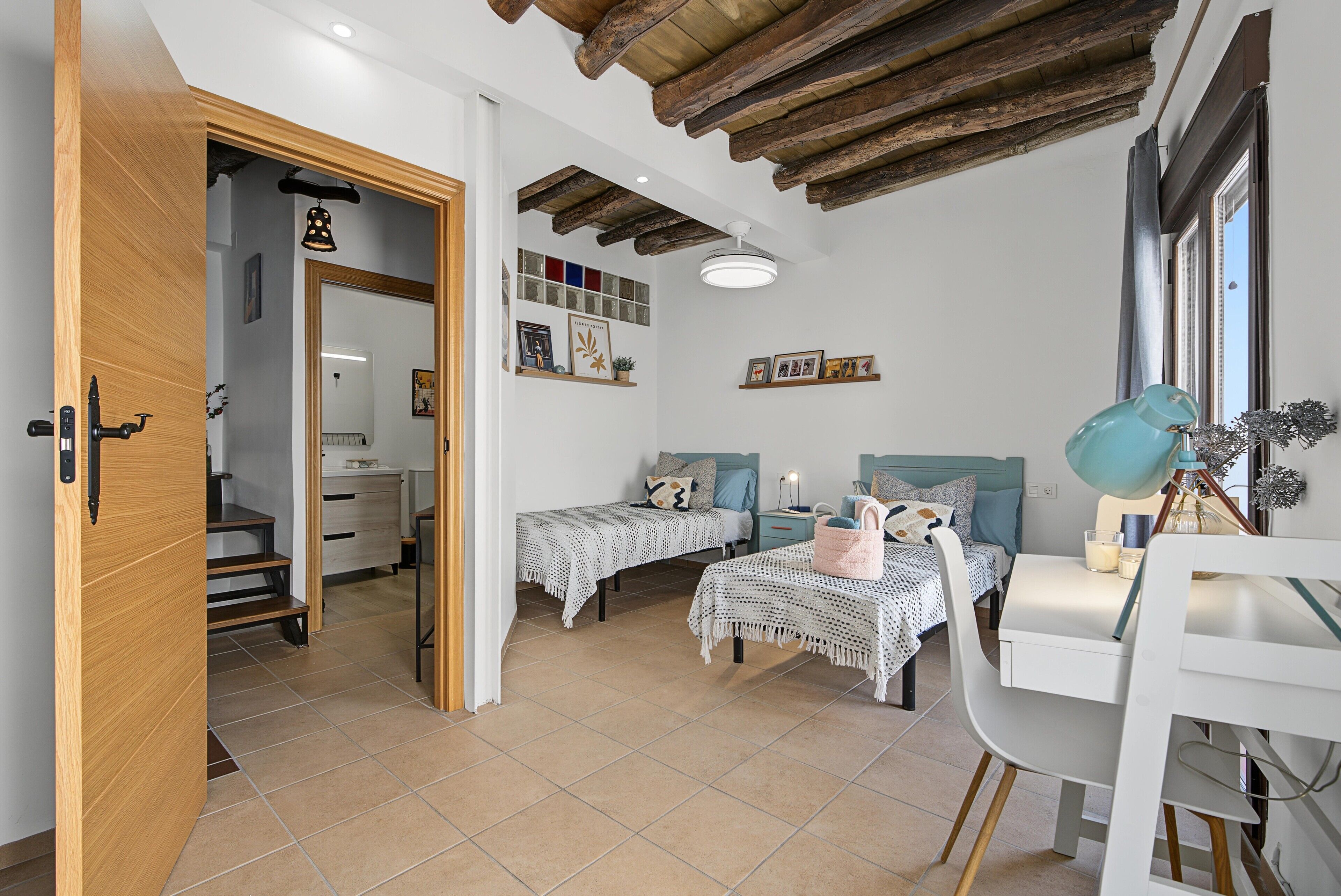 A bedroom with two beds, a desk, and a chair. There is a window with curtains, a ceiling with exposed beams, and a shelf with pictures.